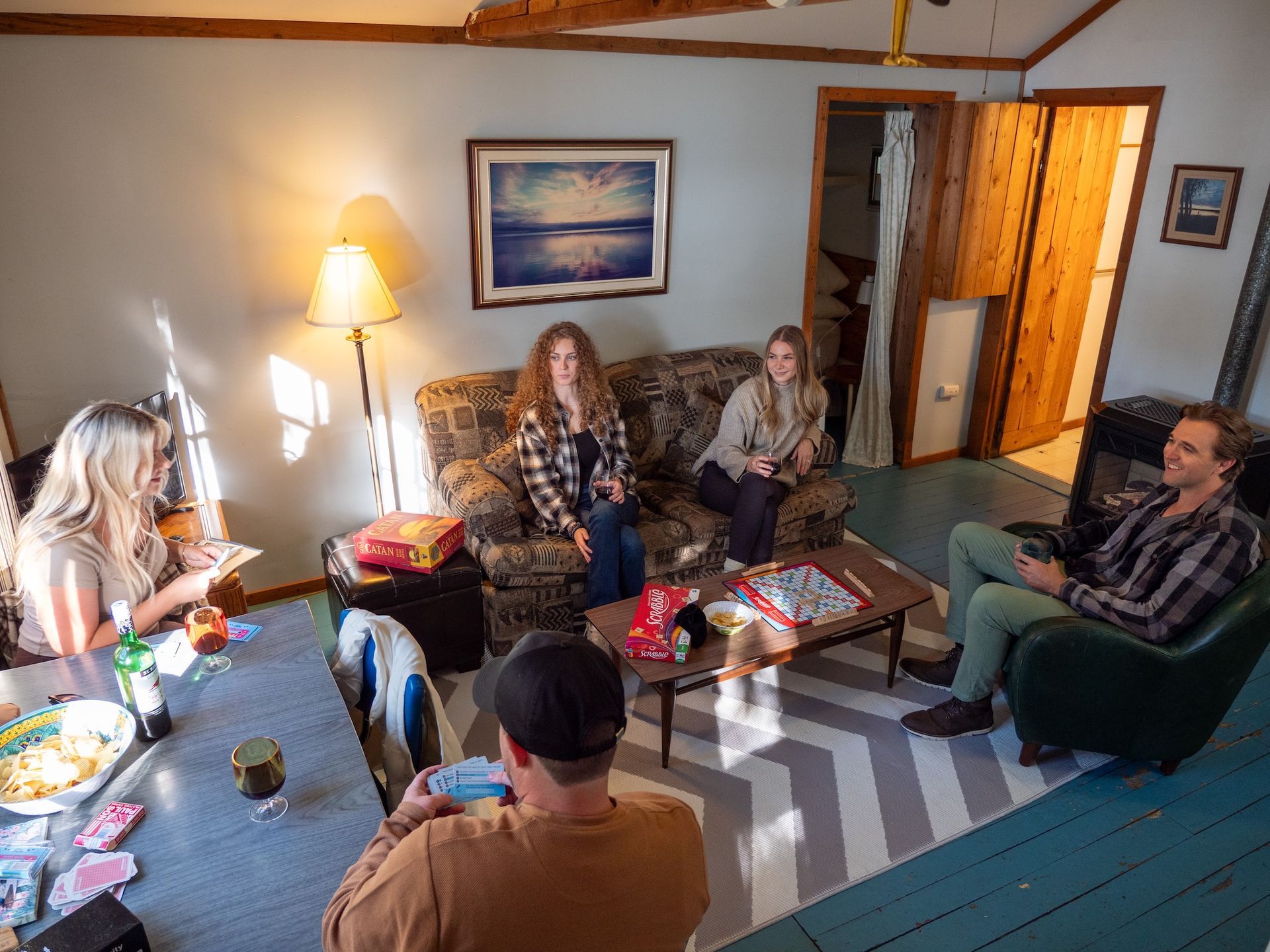 Group of people gathered in living room; playing cards, sitting on a sofas.