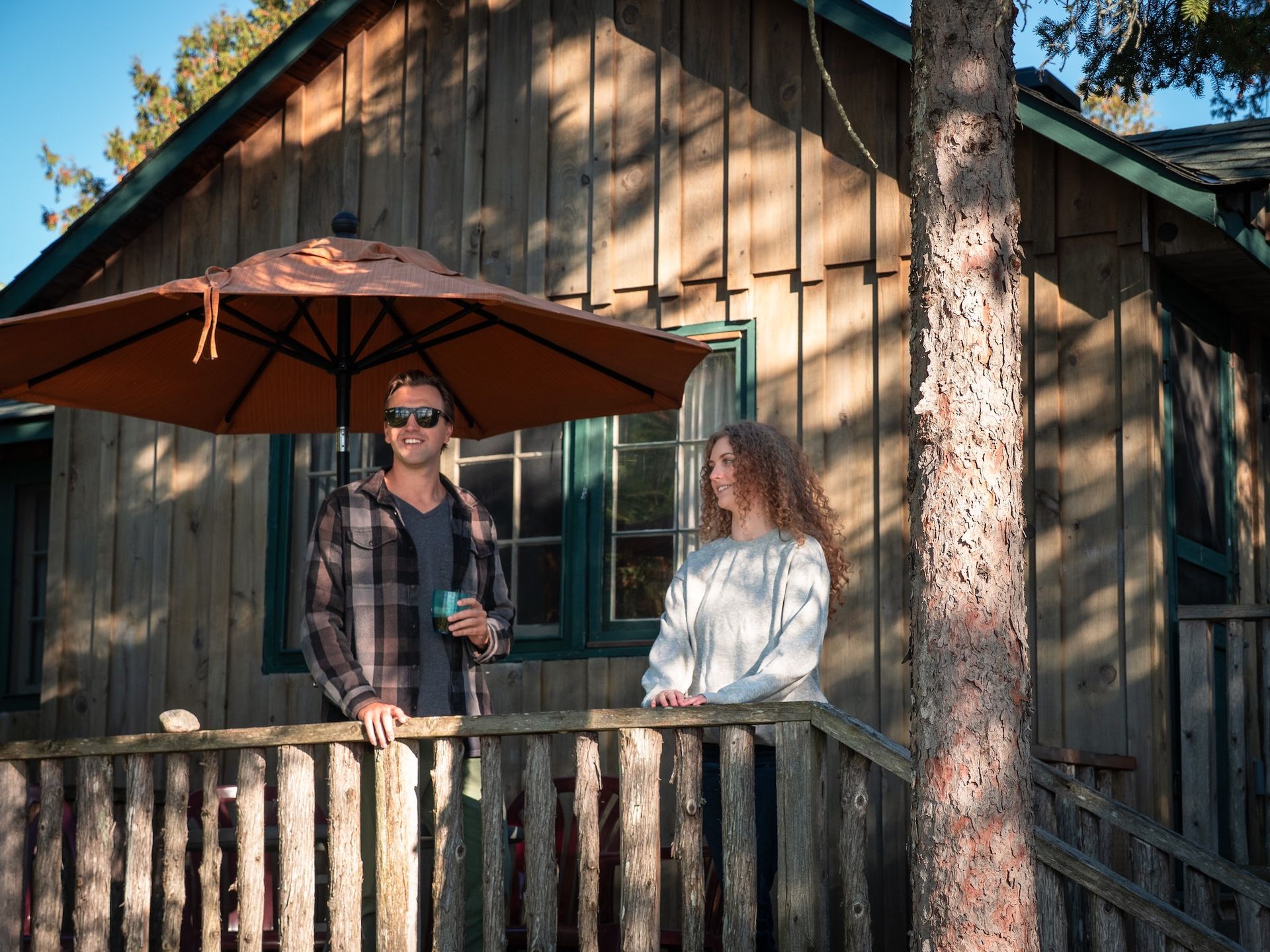 Couple on a cabin porch, shaded by an umbrella. One holds a drink, the other looks on.