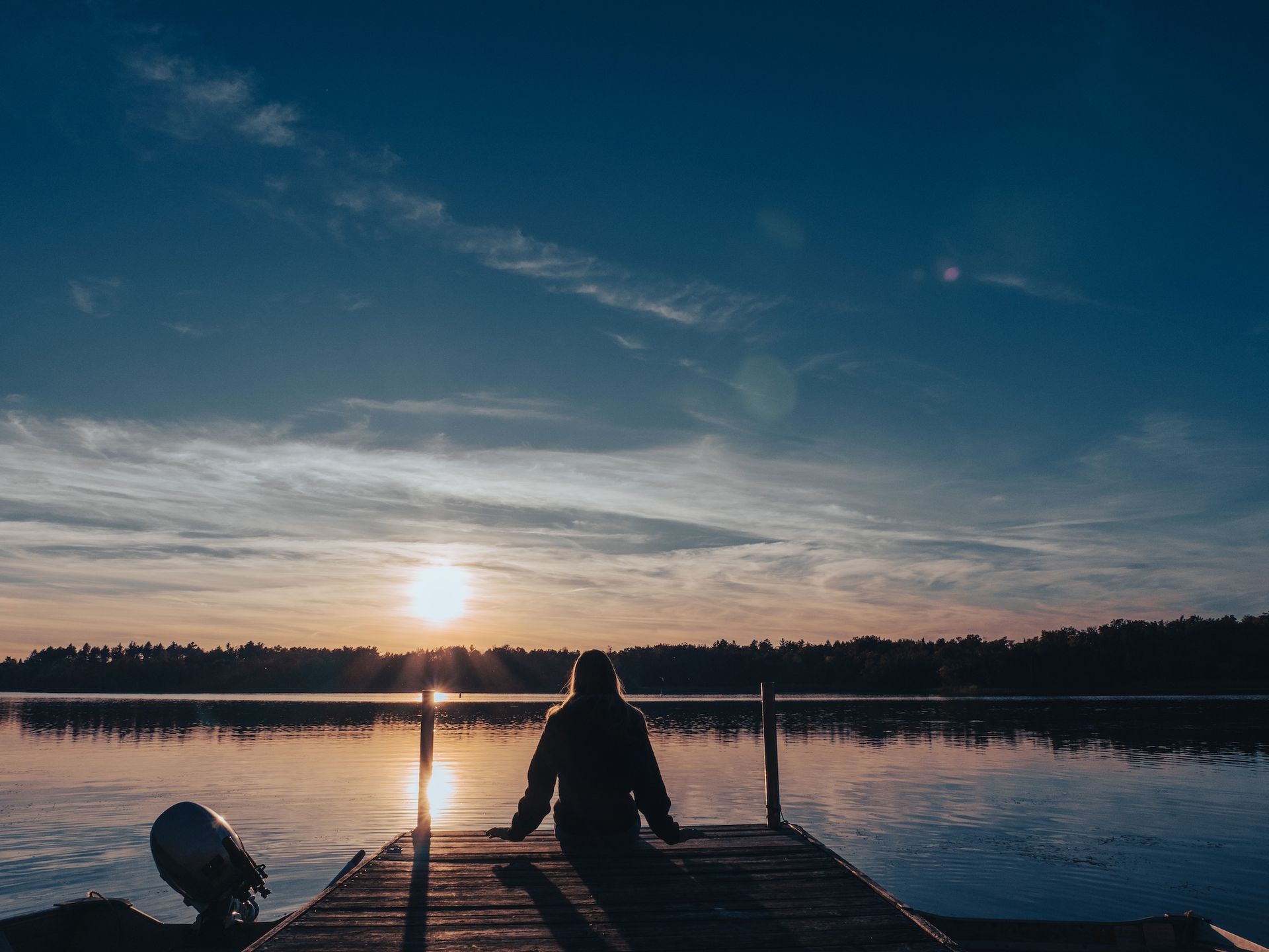 Woman sitting on a wooden dock, facing a sunset over a calm lake.