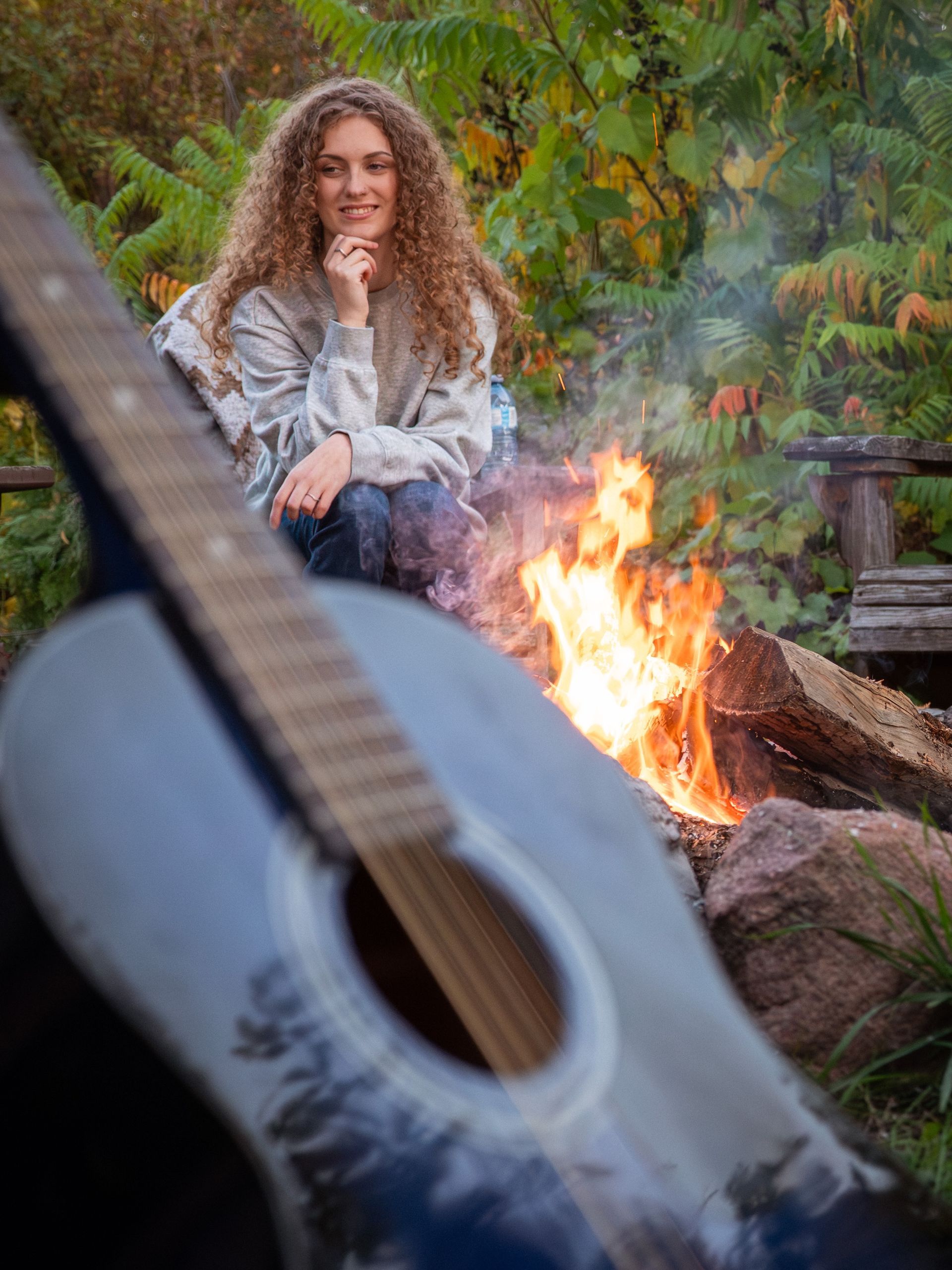 Woman sits by campfire, guitar in foreground. Autumn setting.