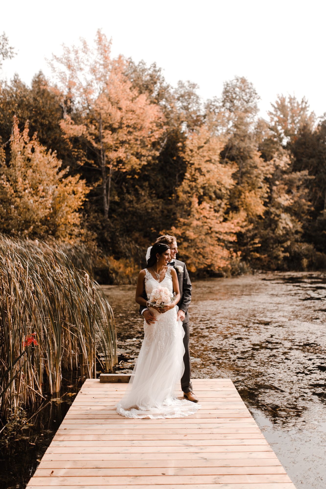 A bride and groom are standing on a wooden dock next to a lake.