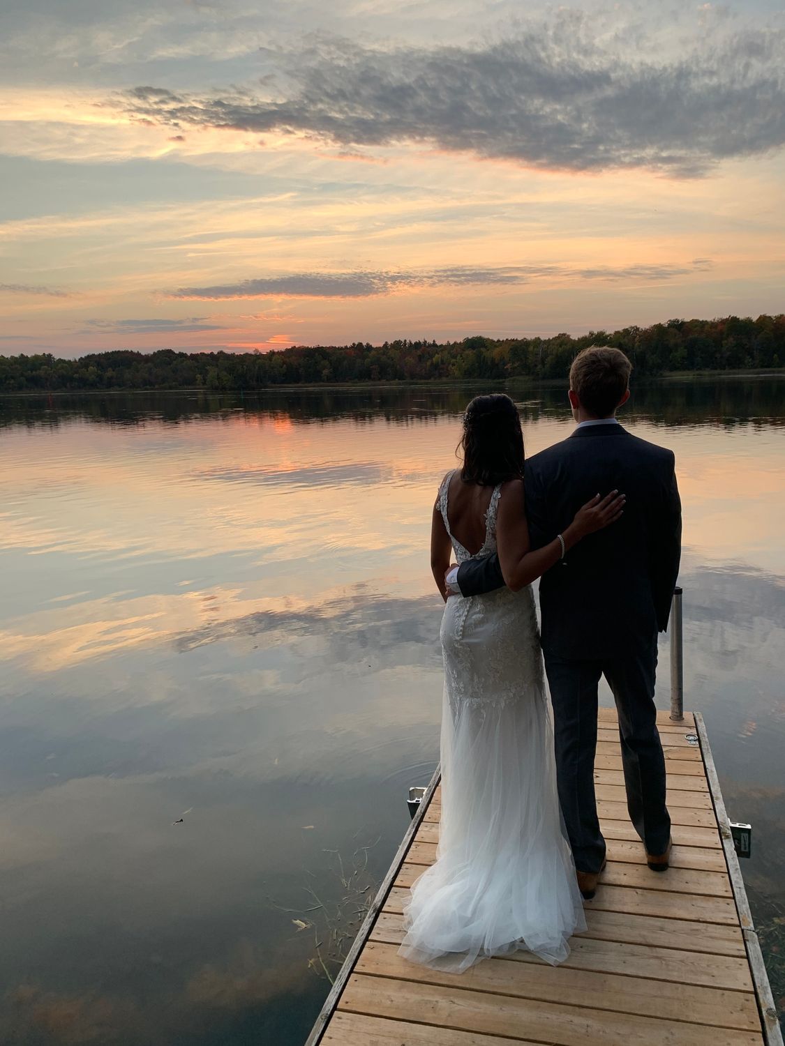 A bride and groom are standing on a dock looking at the sunset over a lake.