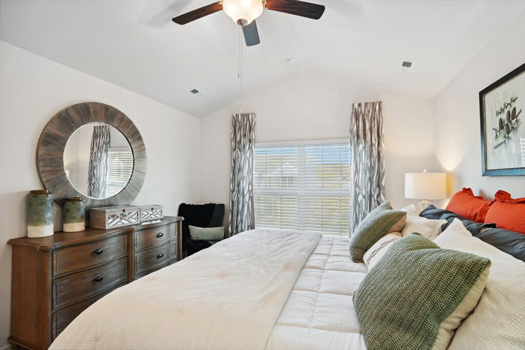 Bedroom with a large bed, wooden dresser, round mirror, and window with curtains.