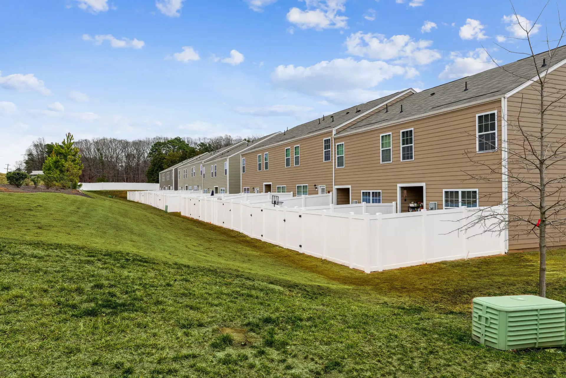 A row of beige townhome buildings with white privacy fences along a grassy slope.