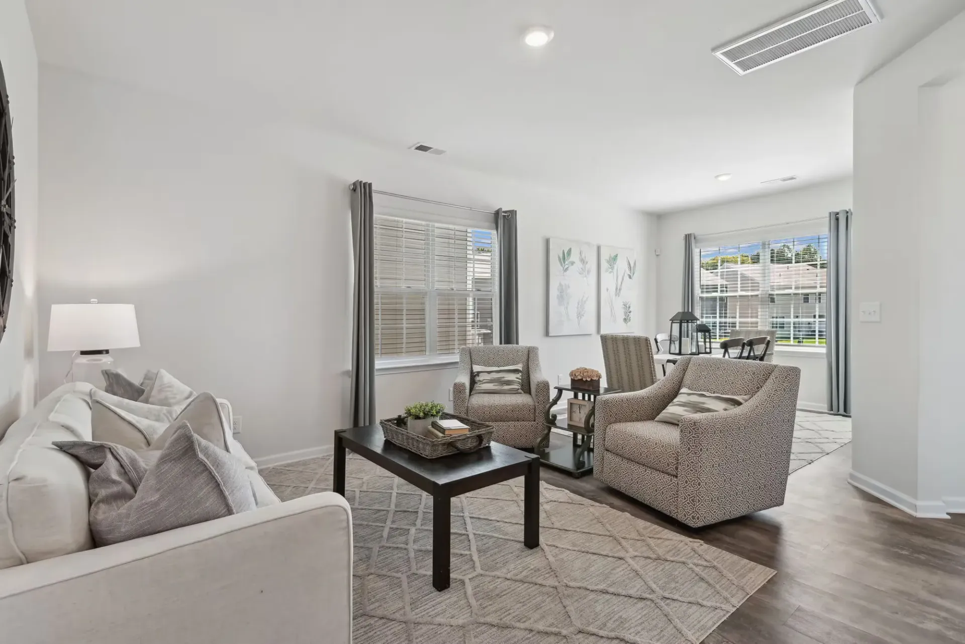 Bright living room with white sofa, two patterned armchairs, a dark coffee table, and large windows with gray curtains.