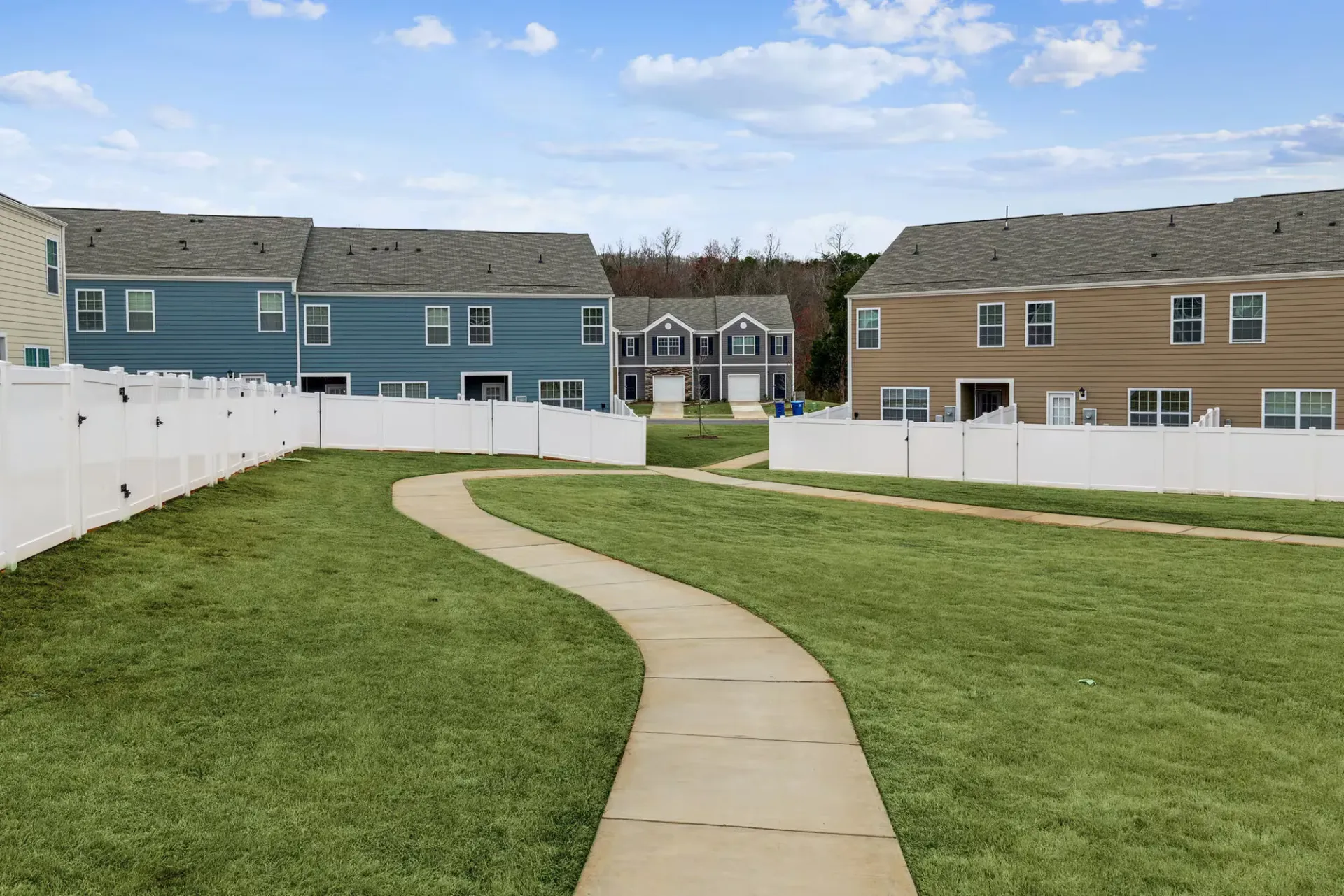 Pathway through green lawn, white fences, and colorful townhomes under a blue sky.