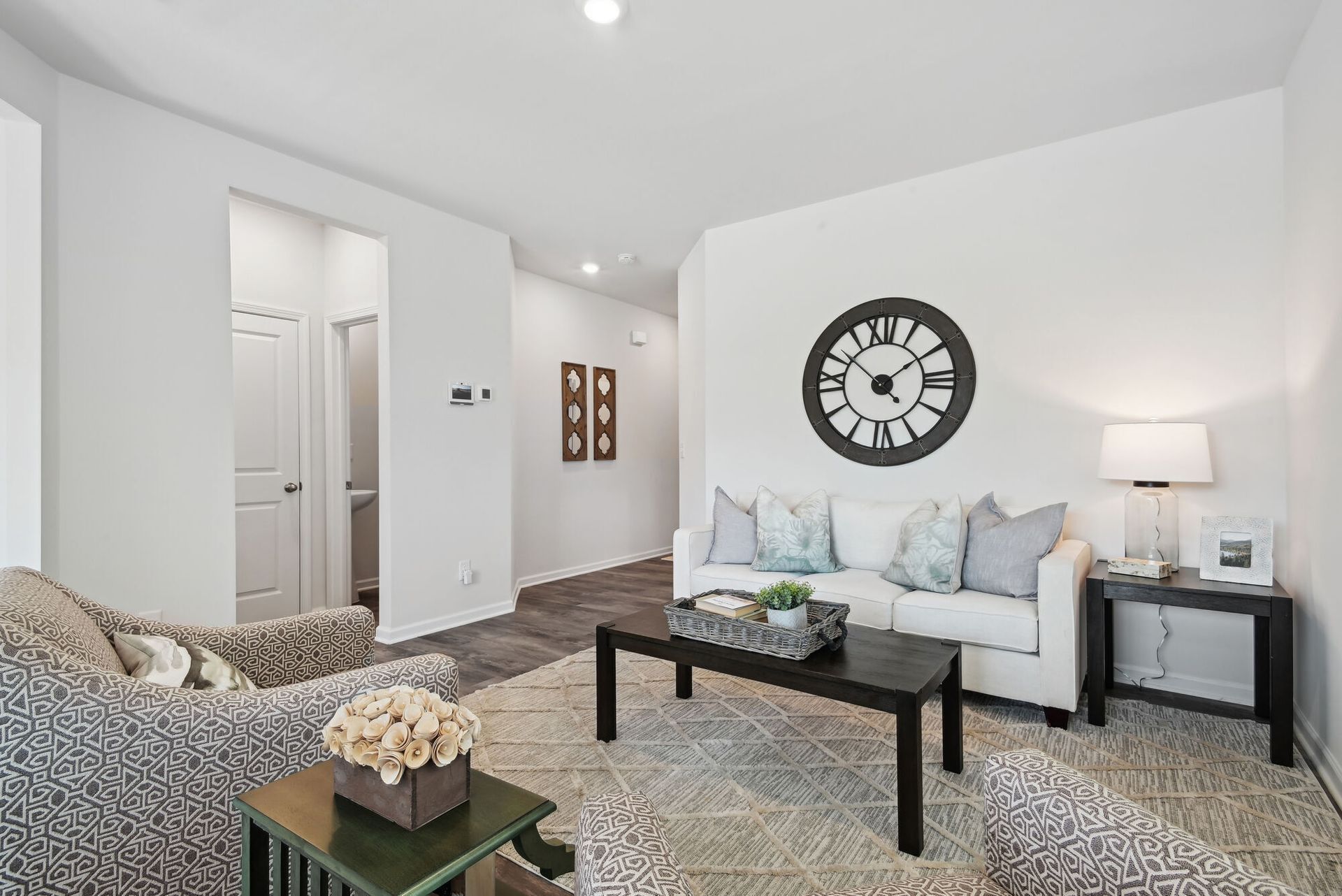 Modern apartment living room with white walls, a gray sofa, patterned chairs, and a large wall clock.