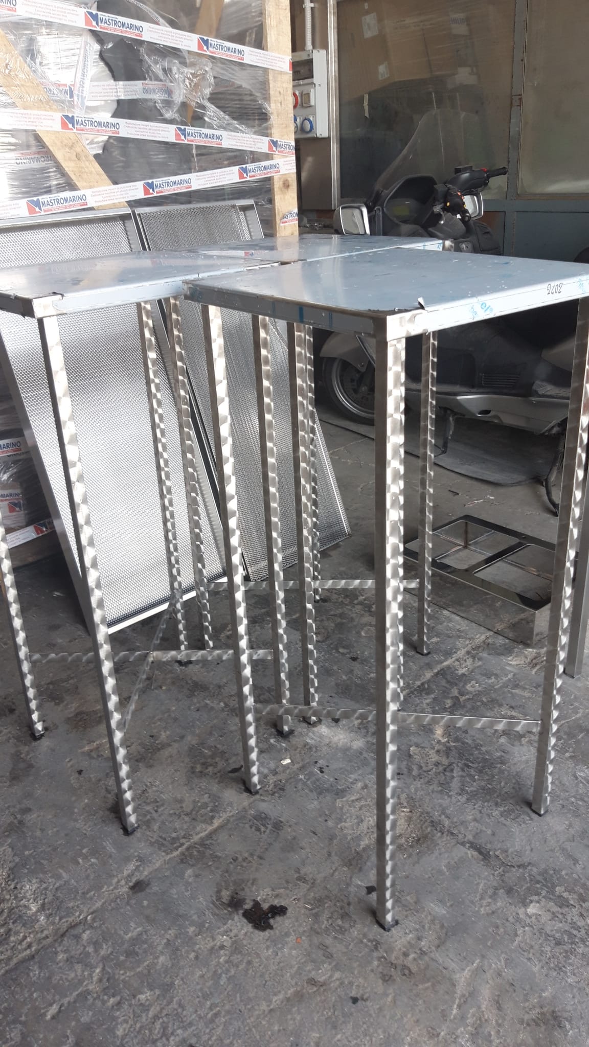 Tavoli in acciaio inox con piani quadrati e gambe sottili, ambiente industriale.