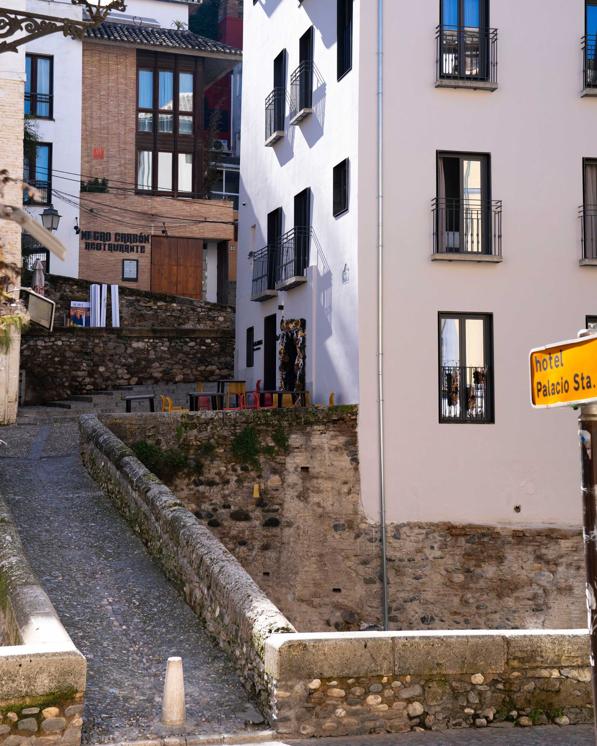 Calle estrecha empedrada entre antiguos edificios blancos y de ladrillo en una ladera, con escalones y balcones.
