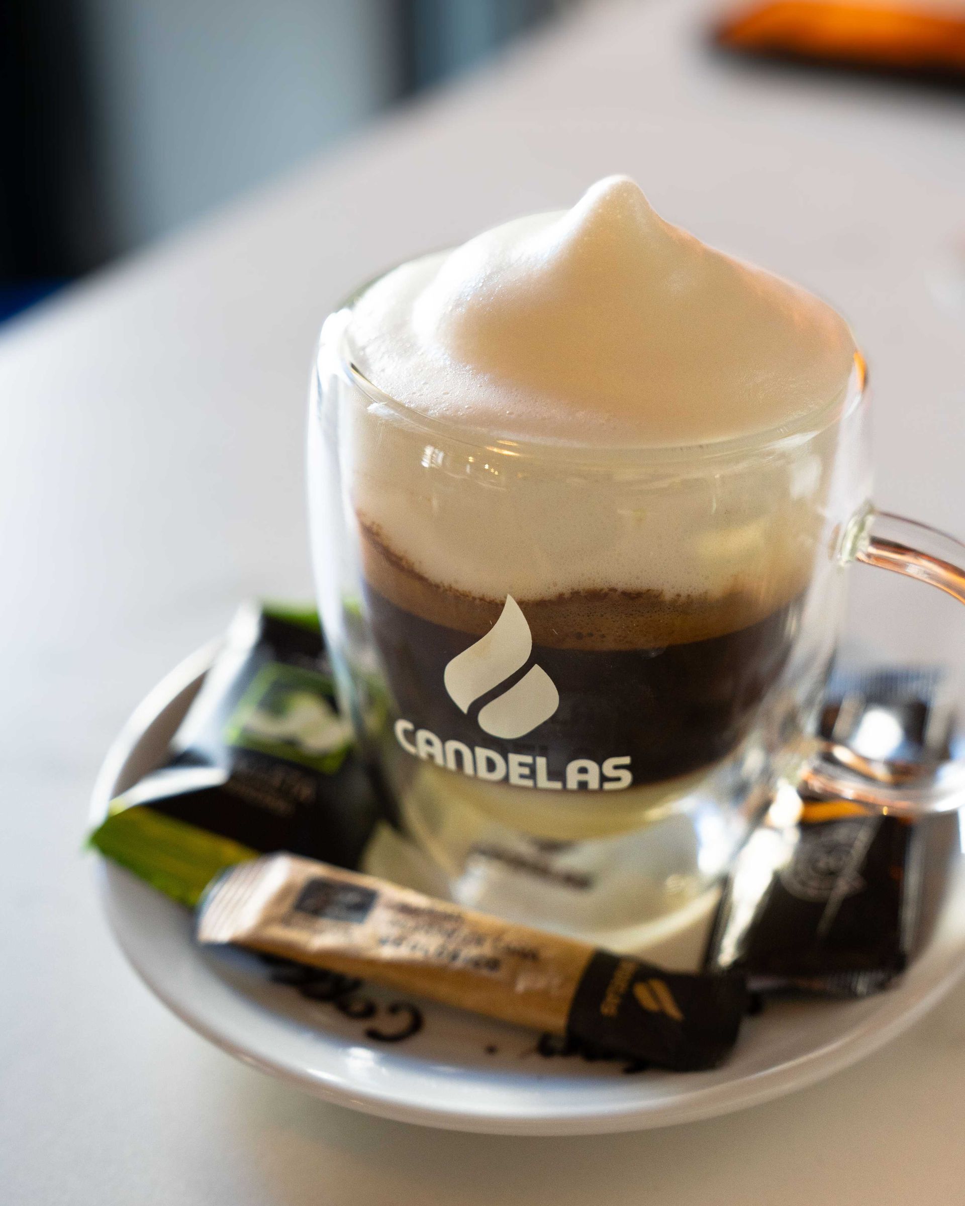 Vaso de capuchino con espuma en un platillo, un caramelo envuelto y el logotipo de la cafetería en la taza.