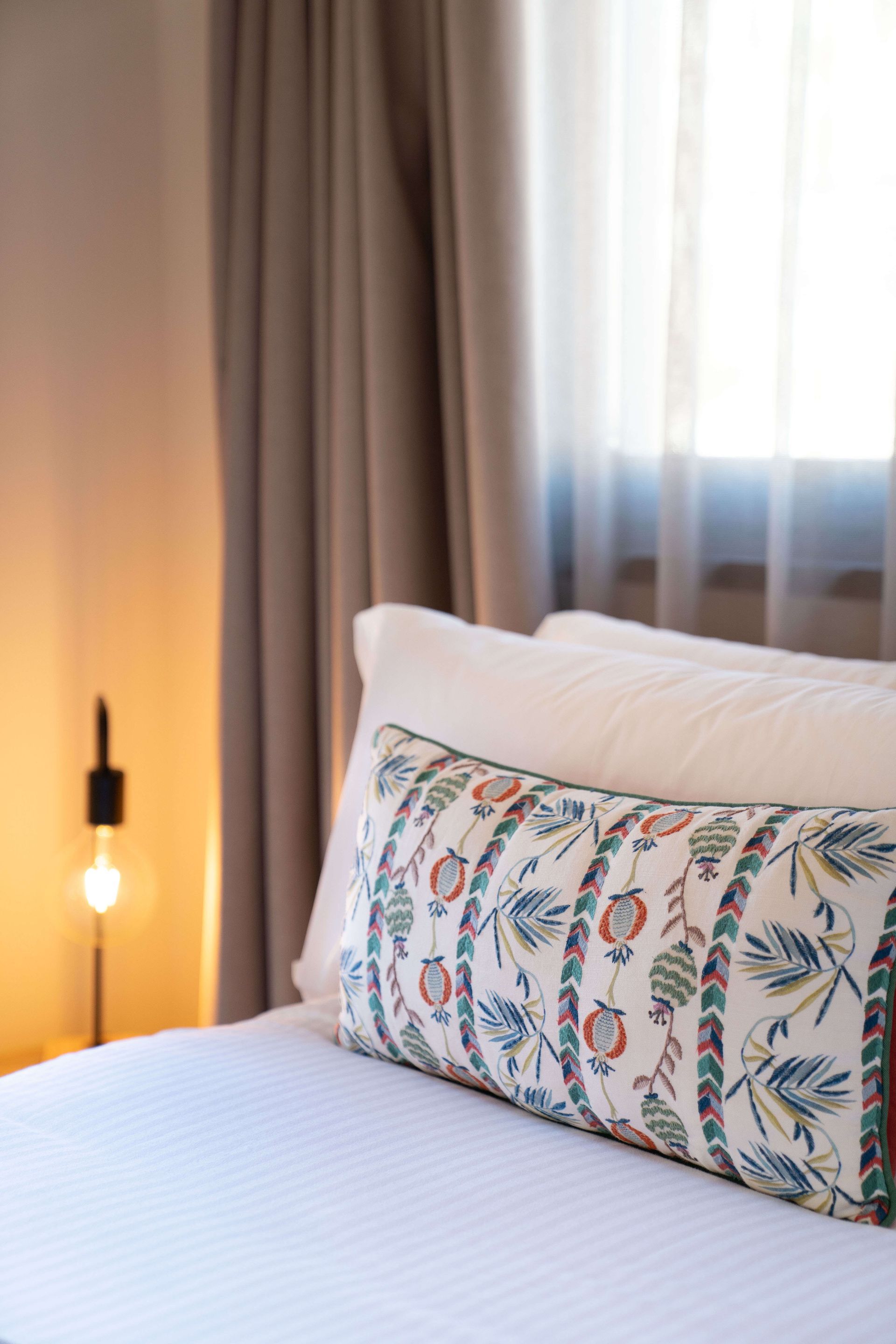 Habitación acogedora con cama, almohada floral, cortinas beige y luz cálida de lámpara cerca de una ventana.