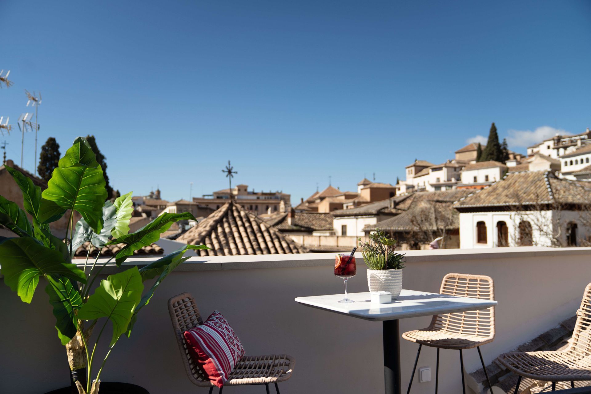 Terraza en la azotea con mesa, sillas, una planta en maceta y vistas a los tejados de terracota bajo un cielo azul.