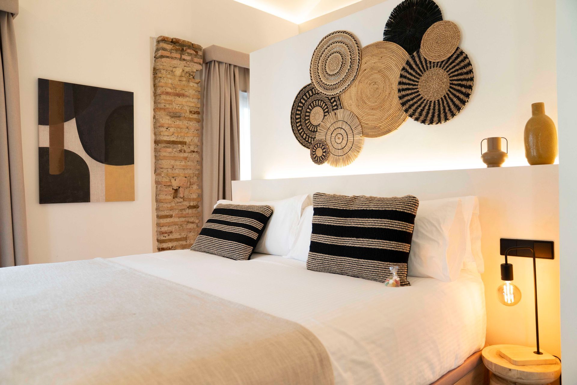Habitación acogedora con cama blanca, almohadas a rayas, cuadro tejido en la pared y cálida iluminación junto a la cama.