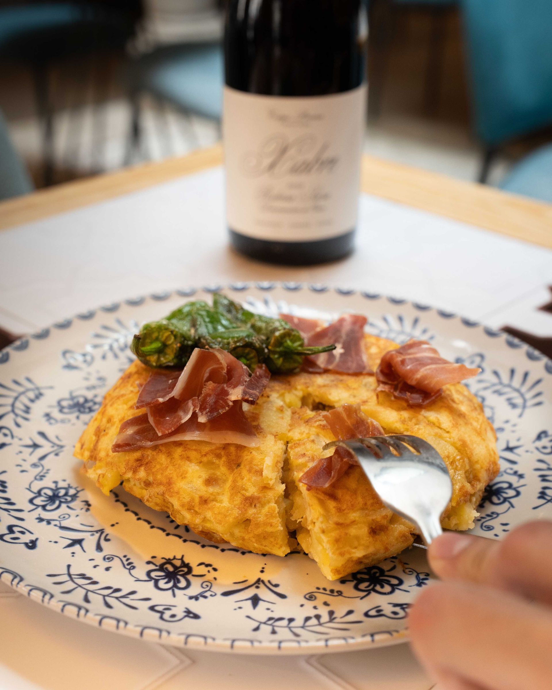Una tortilla esponjosa cubierta con hierbas y jamón serrano en un plato estampado, con un tenedor que la corta.