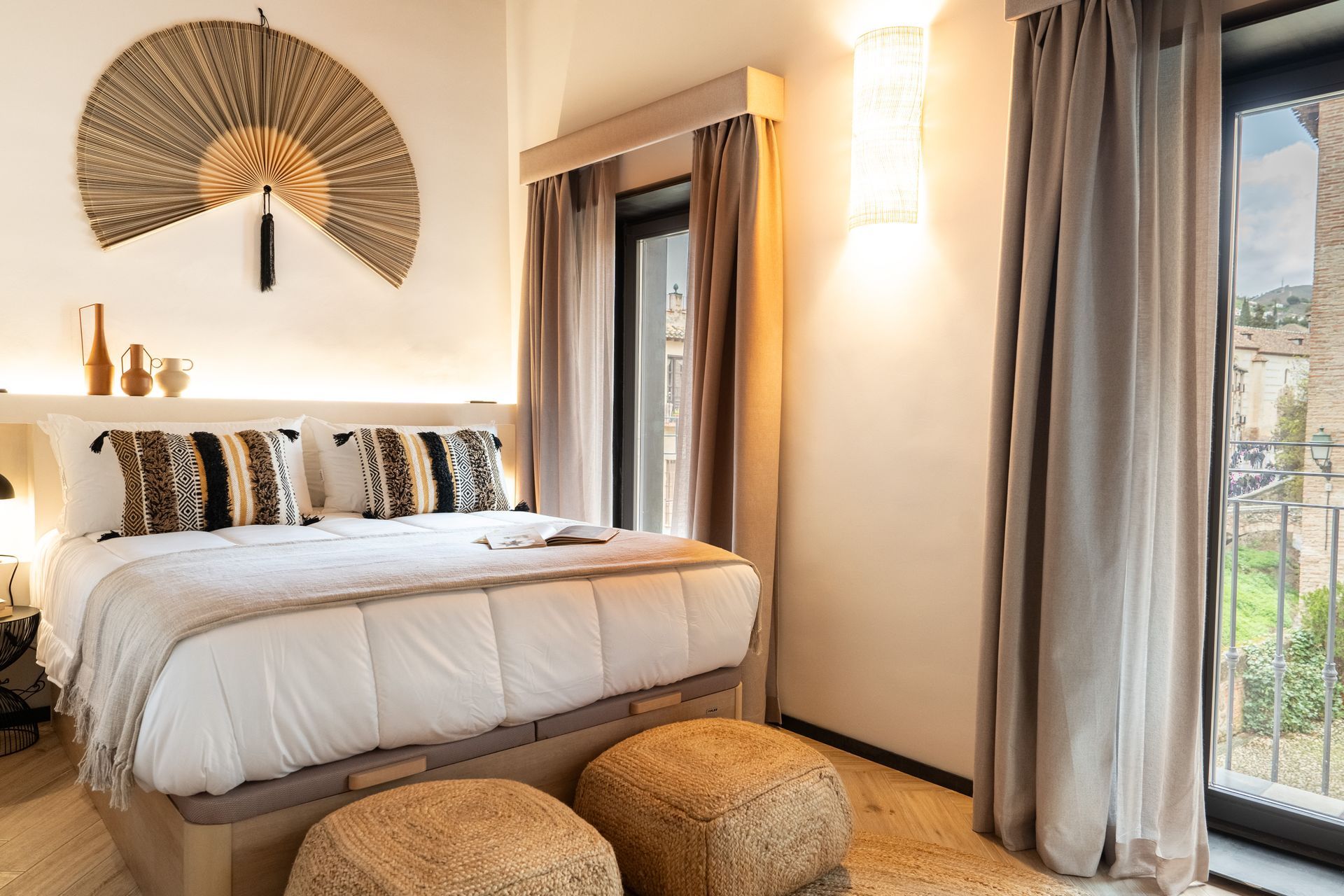 Habitación acogedora con ropa de cama blanca, decoración de pared tejida y una gran ventana con cortinas y vista a la ciudad.