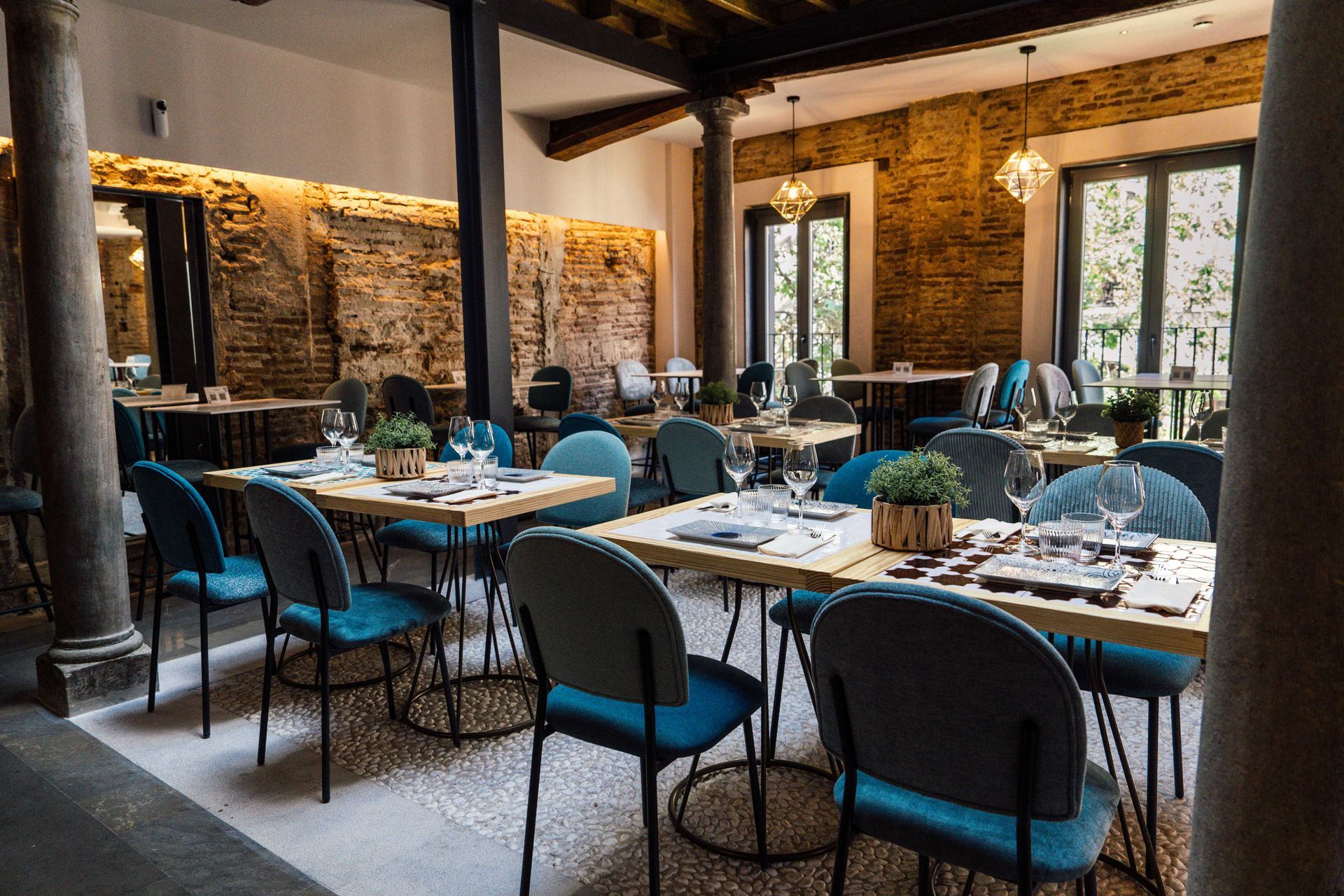 Comedor rústico vacío de un restaurante con sillas azules y mesas dispuestas junto a paredes de ladrillo y grandes ventanales.