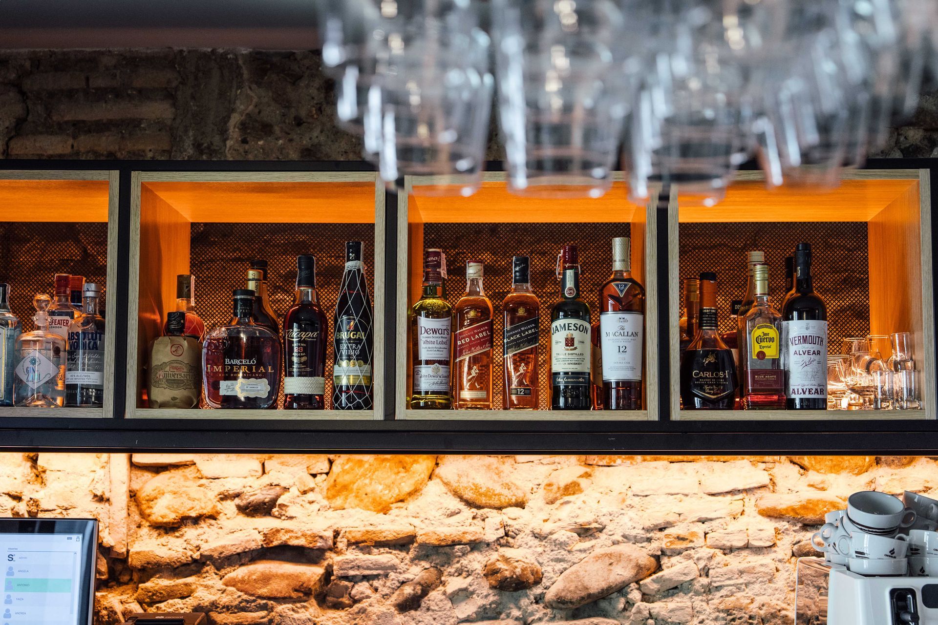 Estanterías con botellas de licor en un bar con iluminación cálida, con una pared de piedra debajo y vasos colgando encima.
