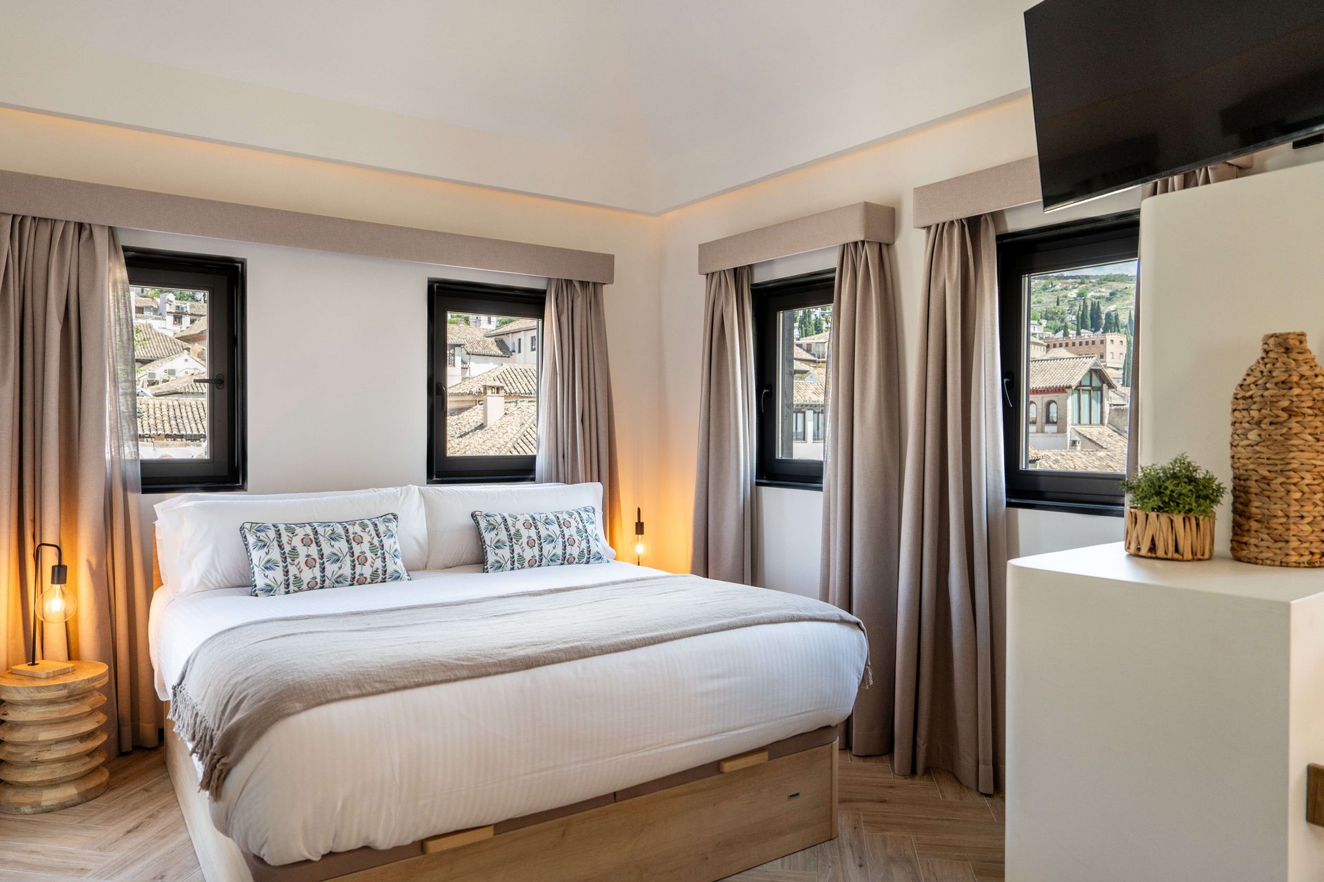Habitación luminosa con una cama grande, decoración neutra y ventanas con vistas a una calle de la ciudad.