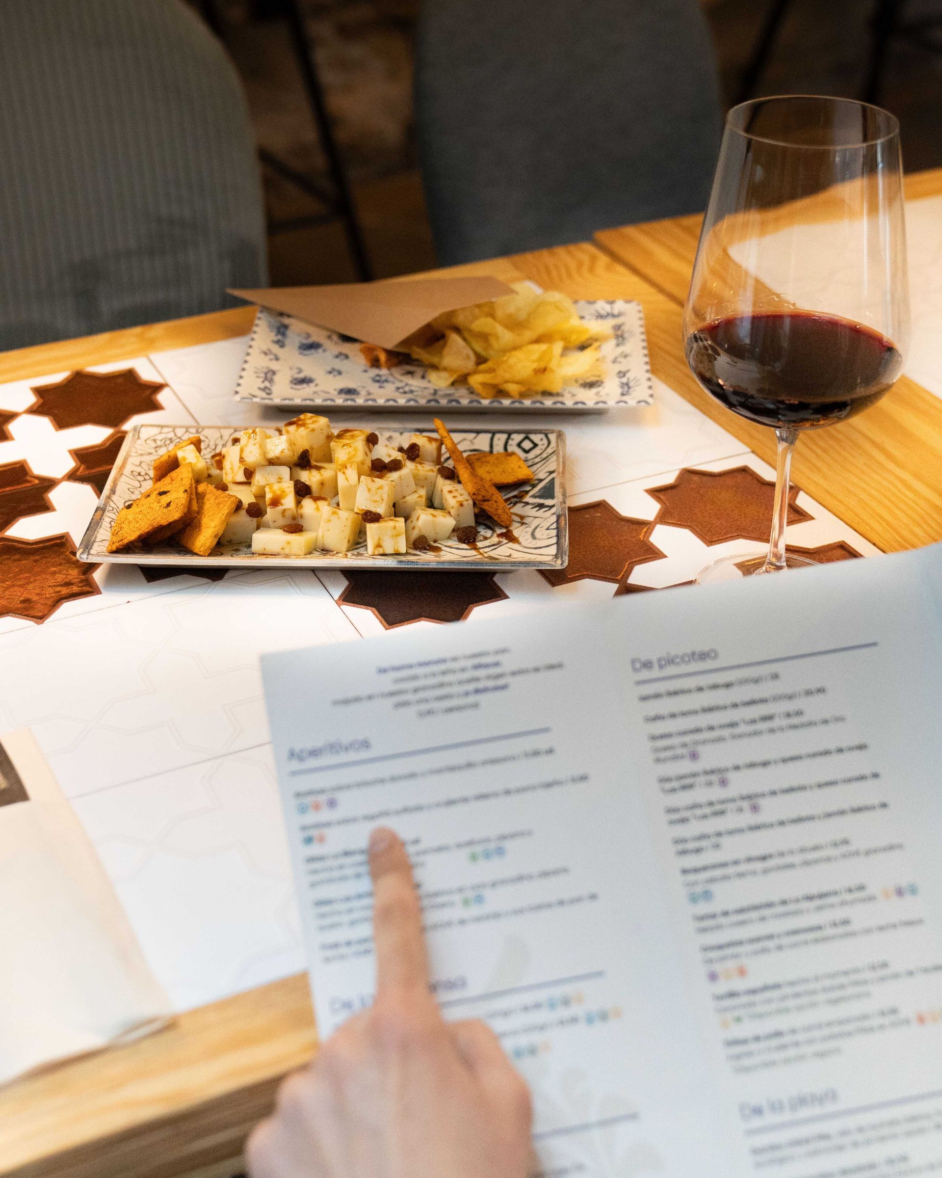Una mano señala el menú impreso junto a los platos de aperitivos y una copa de vino tinto sobre una mesa.