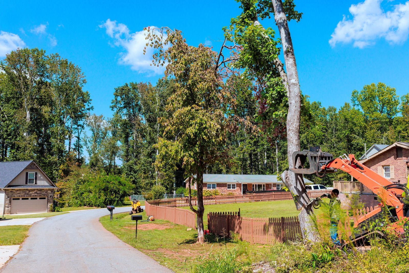 Quiet suburban street with a fenced yard, trees, and houses under a bright blue sky