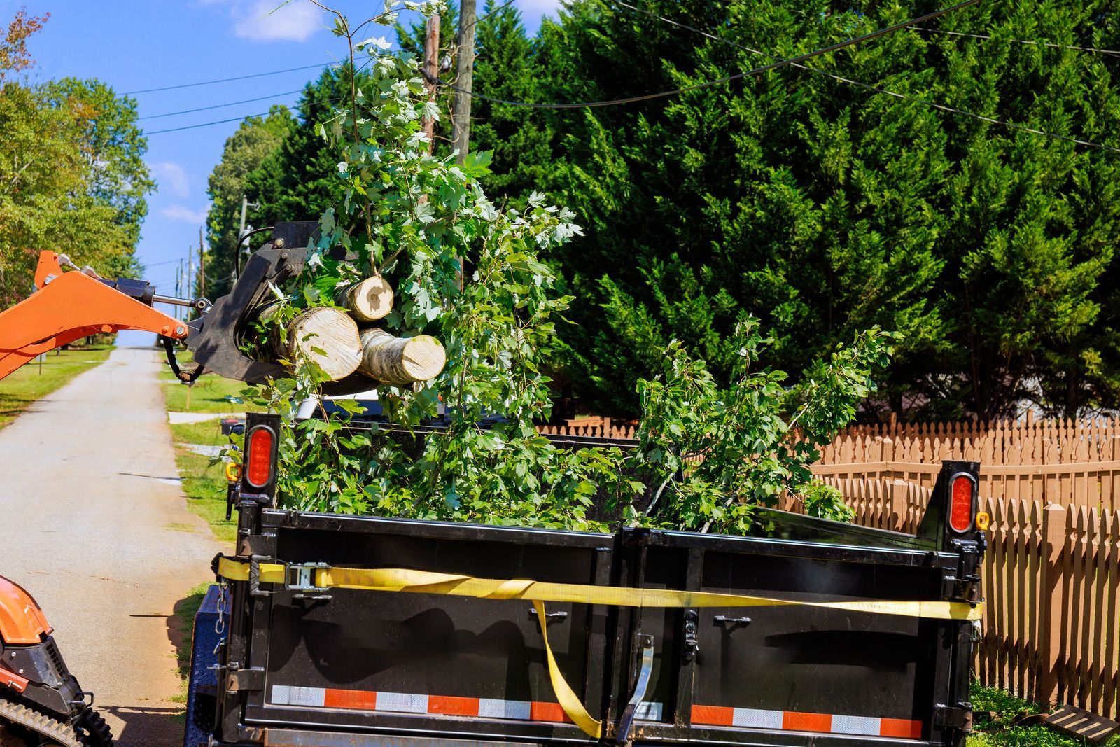 Tree-cutting truck on a narrow road with a worker lifting cut logs near a hedge and fence