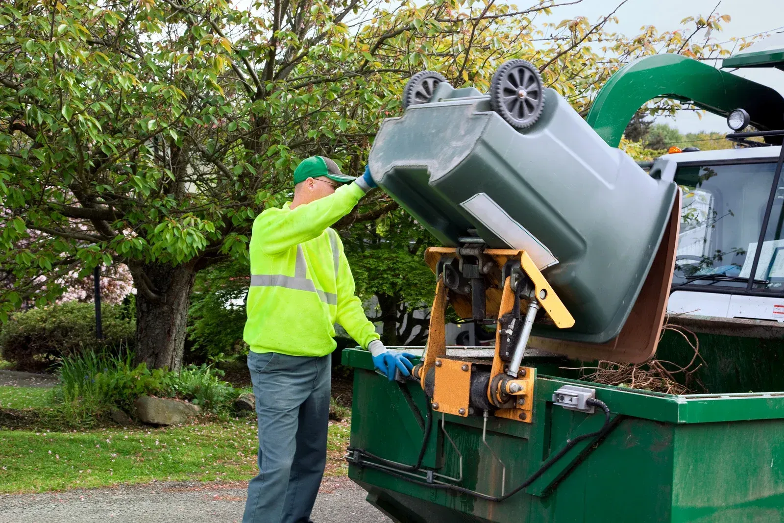 Worker in neon jacket loading a gray trash bin into a green garbage truck outdoors