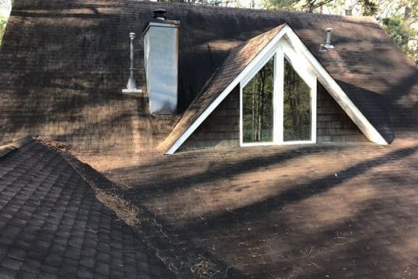 Roof with dark shingles, triangular dormer window, and chimney in sunlight and tree shadows