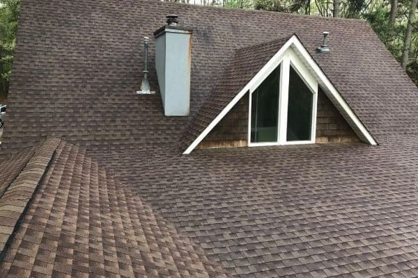 House roof with brown shingles, dormer window, and chimney under trees