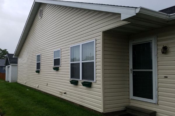 Side of a beige house with vinyl siding, windows, and a screened front door under an overhang