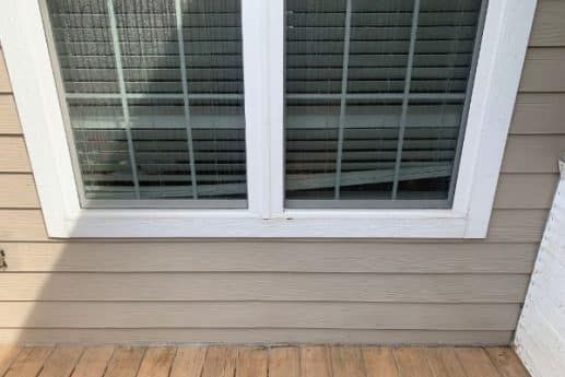 White-framed window with closed blinds above a gray siding wall and wooden deck floor