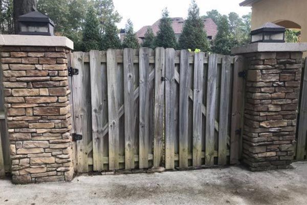 Weathered wooden gate between stone pillars and black lanterns, leading to a driveway.
