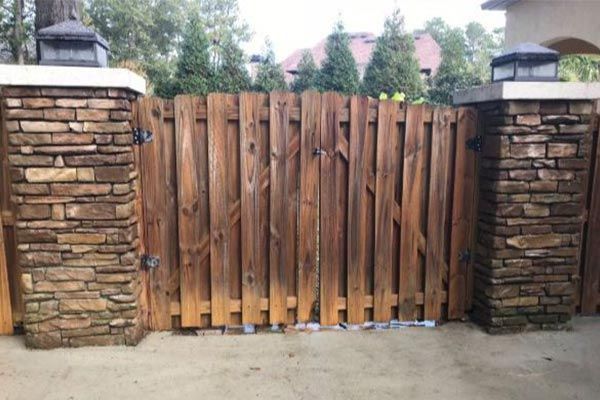 Wooden gate between two stone pillars with black lanterns, in front of a paved driveway