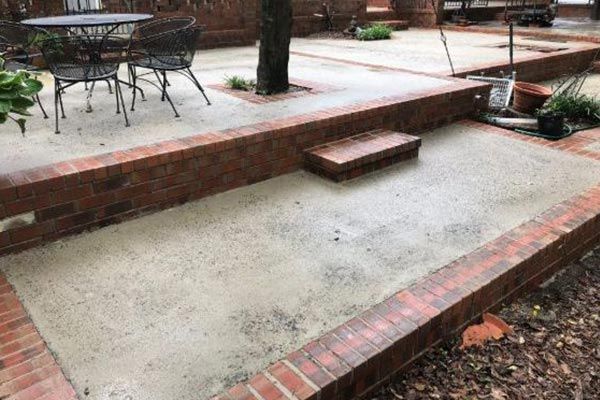 Empty concrete patio with brick edging and a step, beside outdoor furniture and trees.