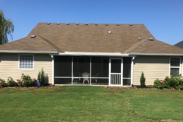 Suburban house with screened back porch, tan siding, brown roof, and green lawn under a clear blue sky