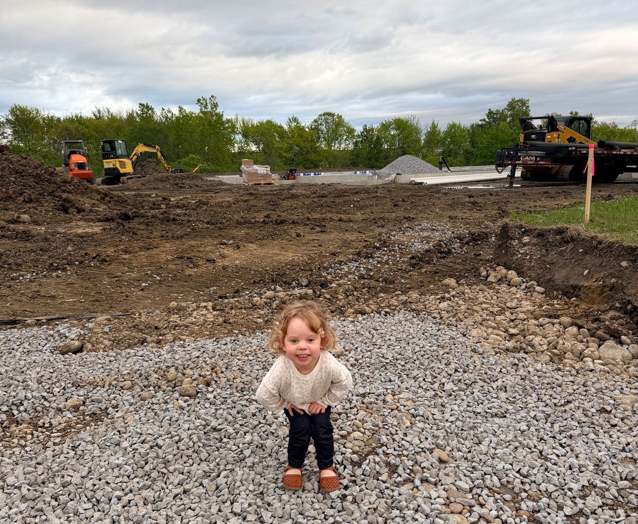 Toddler standing on gravel at a construction site, with heavy equipment and trees in the background
