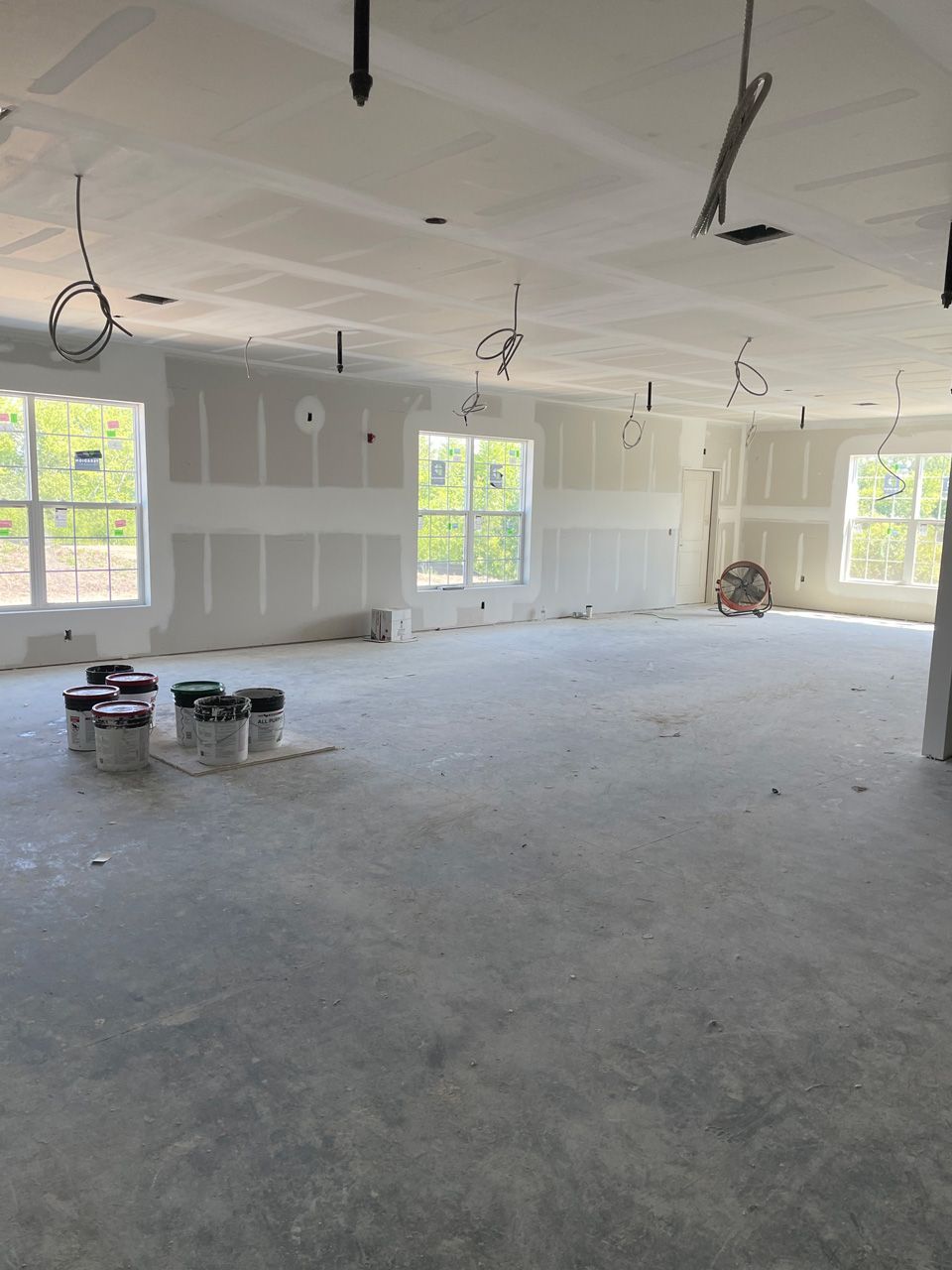 Empty unfinished room with drywall, exposed ceiling wires, and large windows letting in daylight.