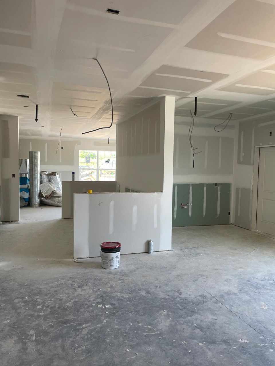 Unfinished interior room with drywall, exposed ceiling wires, and a paint bucket on a dusty floor