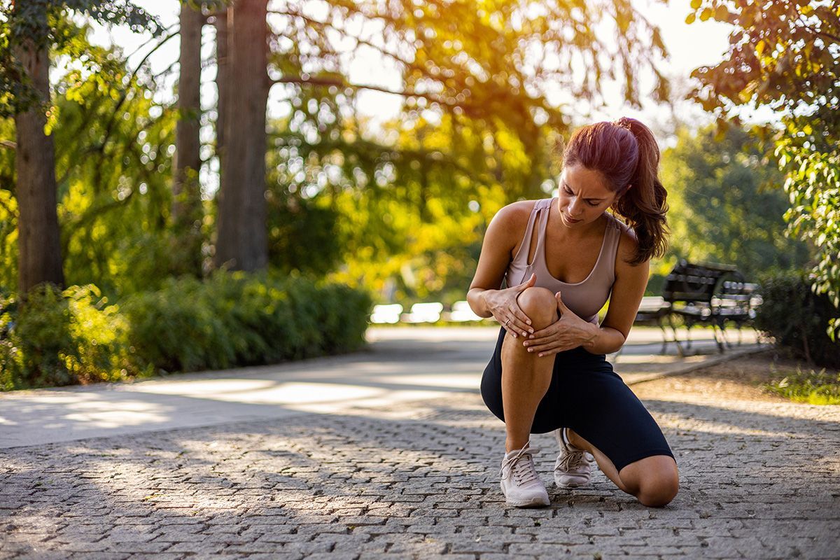 Woman kneeling on a sunlit path outdoors, tying her sneaker in a park with trees and autumn light.