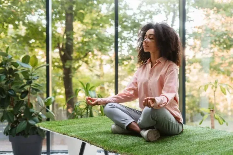 Person meditating cross-legged on a grassy rooftop beside plants and large windows.