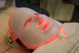 A white facial LED mask glowing red, resting on a person’s face during a spa treatment.