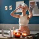 Person sitting on a couch applying a towel to their head, with candles and bottles on a coffee table in a blue living room.