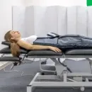 Person lying on a padded exam table in a clinic, with a white privacy screen behind and medical equipment below.