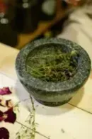 Mortar and pestle with crushed herbs on a table, with scattered rose petals and a hand nearby