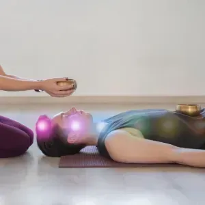 Two people practicing yoga; one kneels holding a singing bowl over the other lying on a mat with crystal bowls nearby