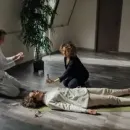 Two people kneel beside a woman lying on the floor in a bright room, as one appears to check her breathing.