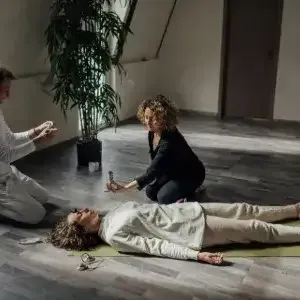 Two people kneel beside a woman lying on the floor in a bright room, as one appears to check her breathing.