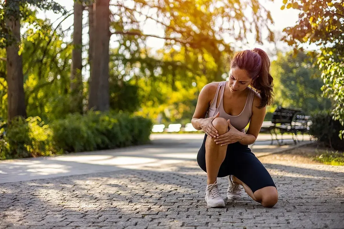 Woman kneeling on a sunlit path outdoors, tying her sneaker in a park with trees and autumn light.