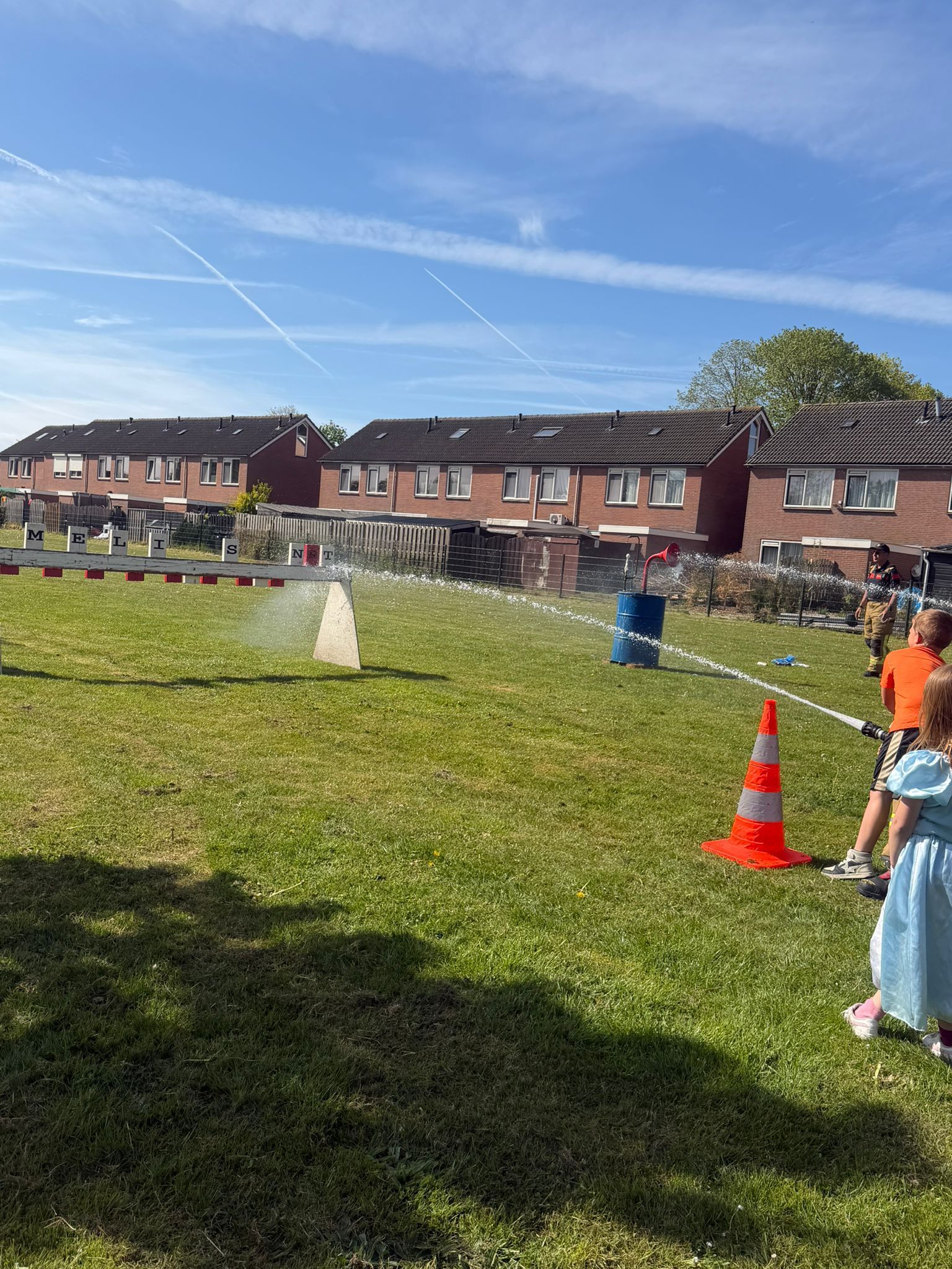 Kinderen spelen bij een sproeier op een zonnig grasveld met oranje kegels en huizen op de achtergrond.