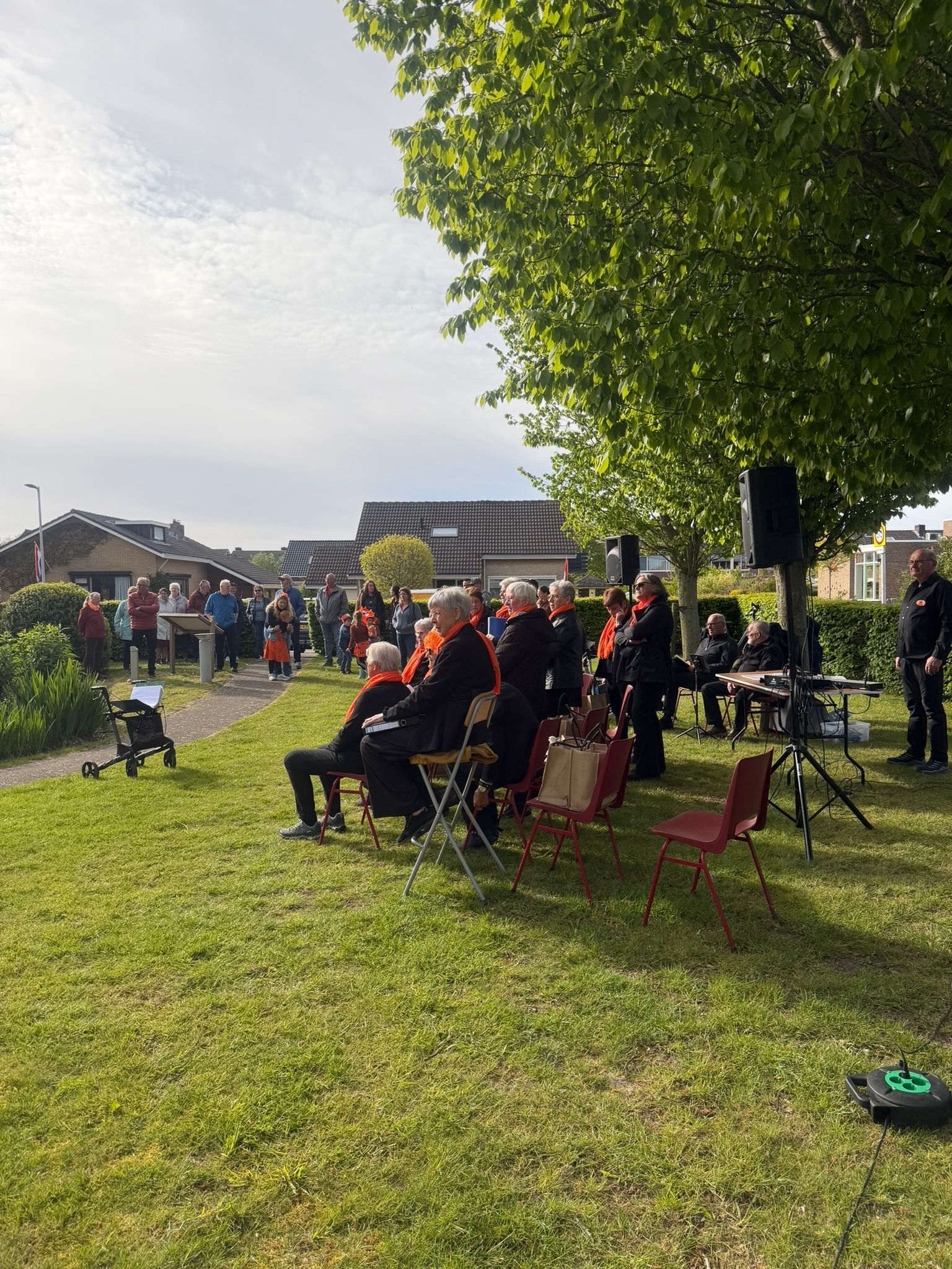 Mensen zitten op stoelen in een zonnig park, met op de achtergrond een menigte en tenten.