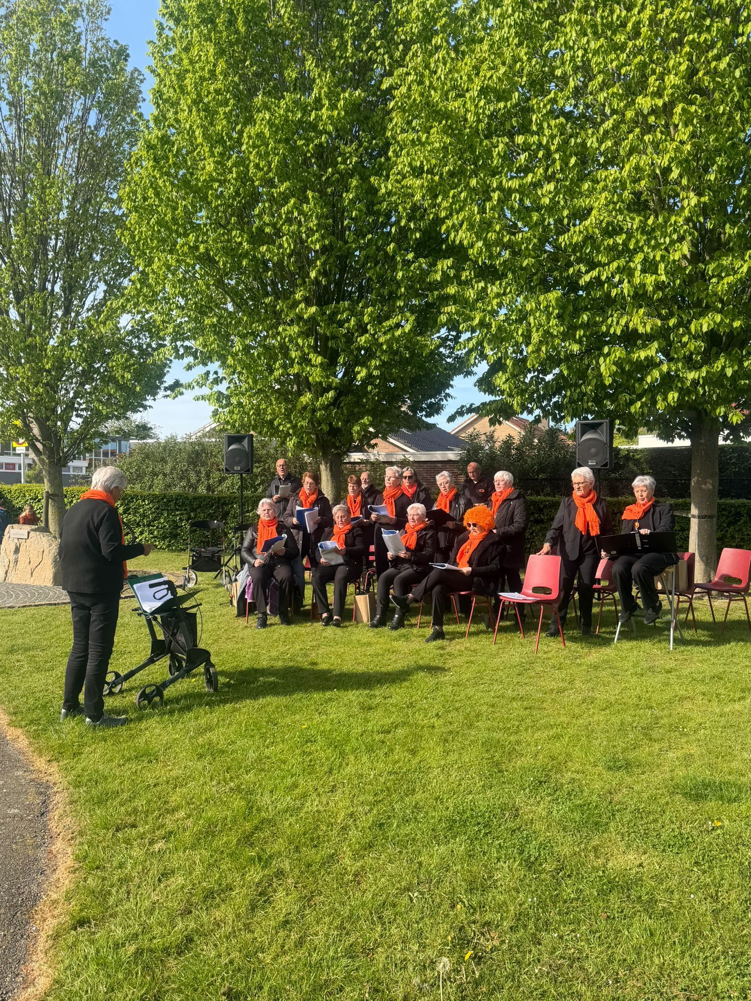 Een koor in oranje hesjes zit buiten onder de bomen, met een dirigent links van hen.